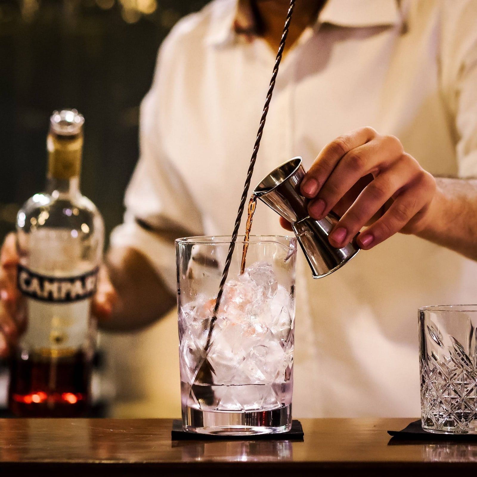 A bartender pouring Campari from a jigger into a mixing glass of ice, the bottle beside it