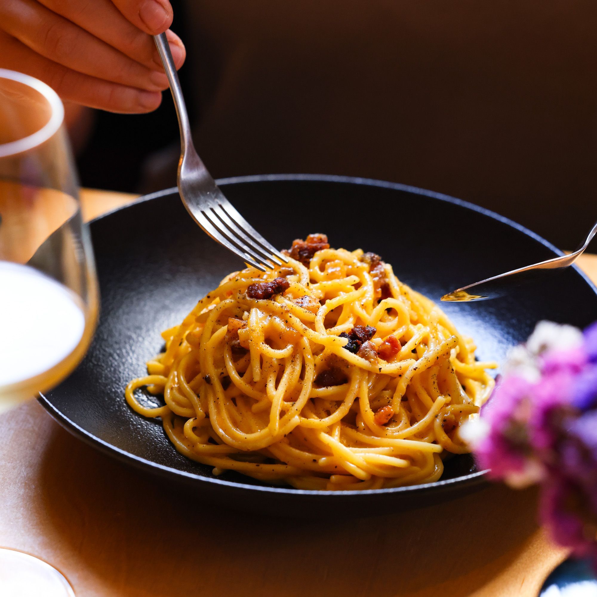 Spaghetti alla carbonara con guanciale croccante in una ciotola nera, accanto un bicchiere di vino bianco