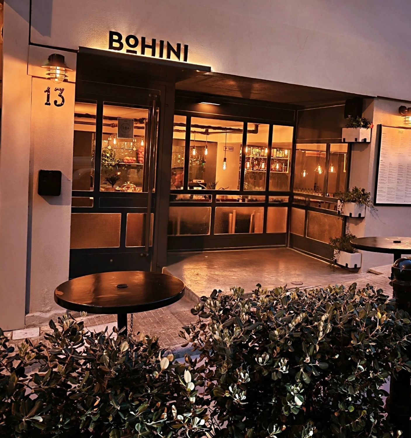L'ingresso di Bohini al tramonto: l'insegna BOHINI illuminata sopra il numero 13, luci calde dietro le vetrate e tavolini all'aperto