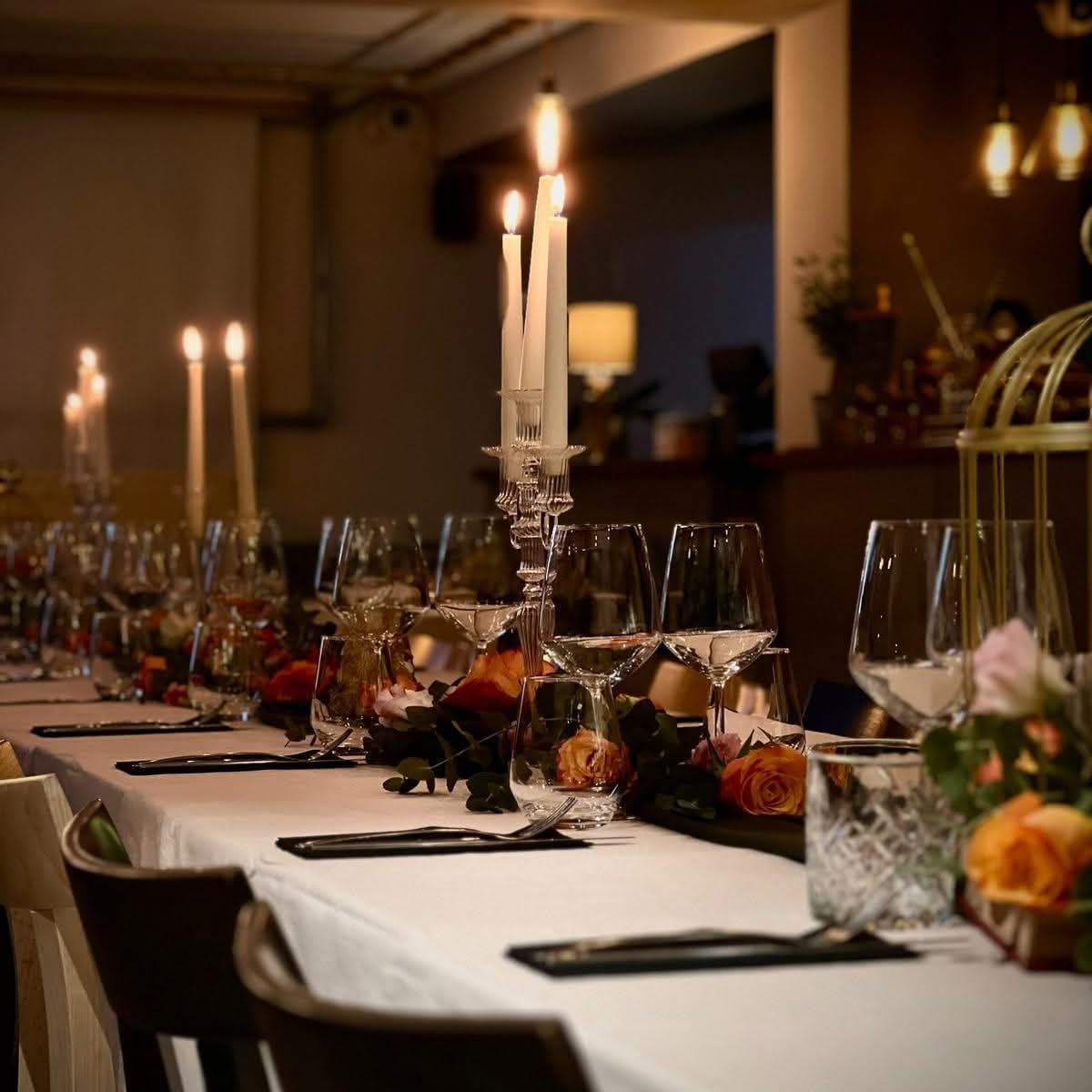 A long table set for a private dinner, with candles, orange roses and wine glasses on a white tablecloth